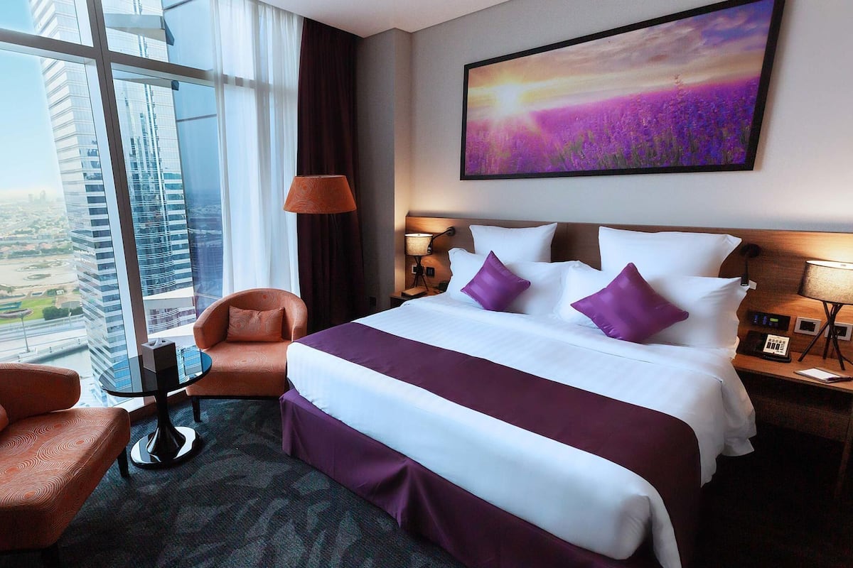 Ein Hotelzimmer mit einem großen Bett, einem Sessel, einem kleinen Tisch und Blick auf eine Stadtlandschaft durch große Fenster.