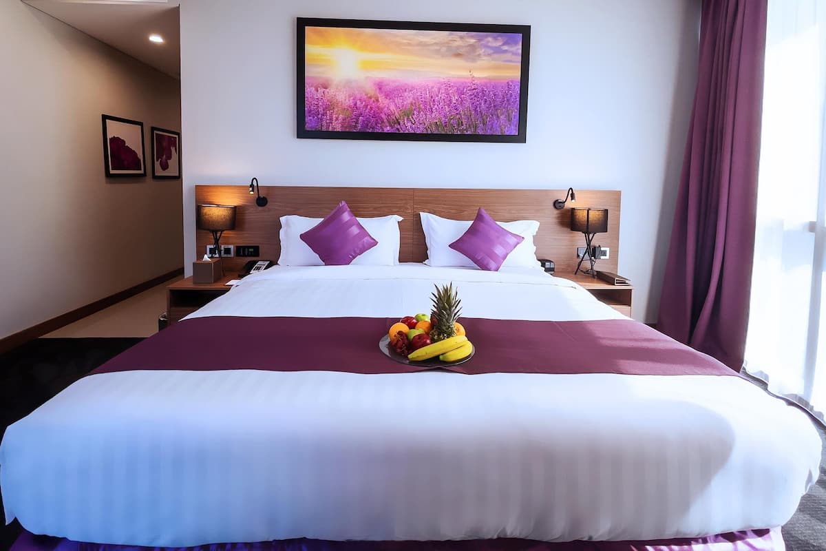 Ein Hotelzimmer mit einem großen Bett, violetten Kissen, einer Obstschale auf dem Bett, einem Gemälde einer Untergangssonne und einem Nachttisch mit einer Lampe.