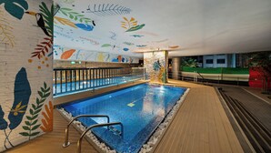 Une piscine intérieure dotée d’une terrasse en bois, entourée de murs ornés de fresques représentant des feuilles et des oiseaux.