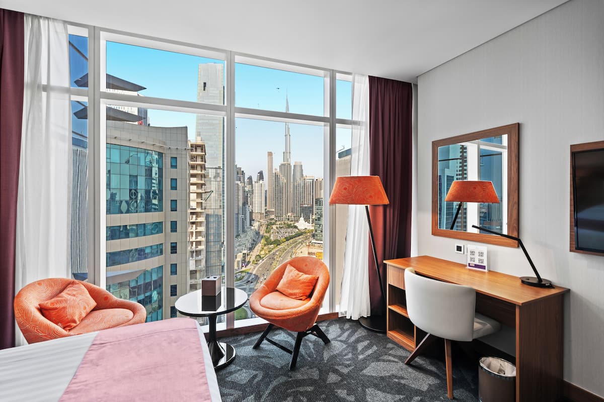 A hotel room with a large window, a desk, a chair, and a bed.