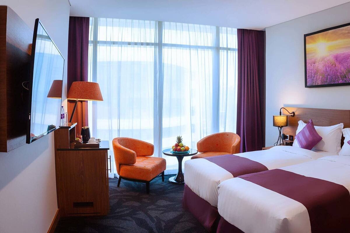 Ein Hotelzimmer mit zwei Betten, einem Schreibtisch, einem Stuhl, einem kleinen Tisch mit Obst und einem großen Fenster mit Vorhängen.
