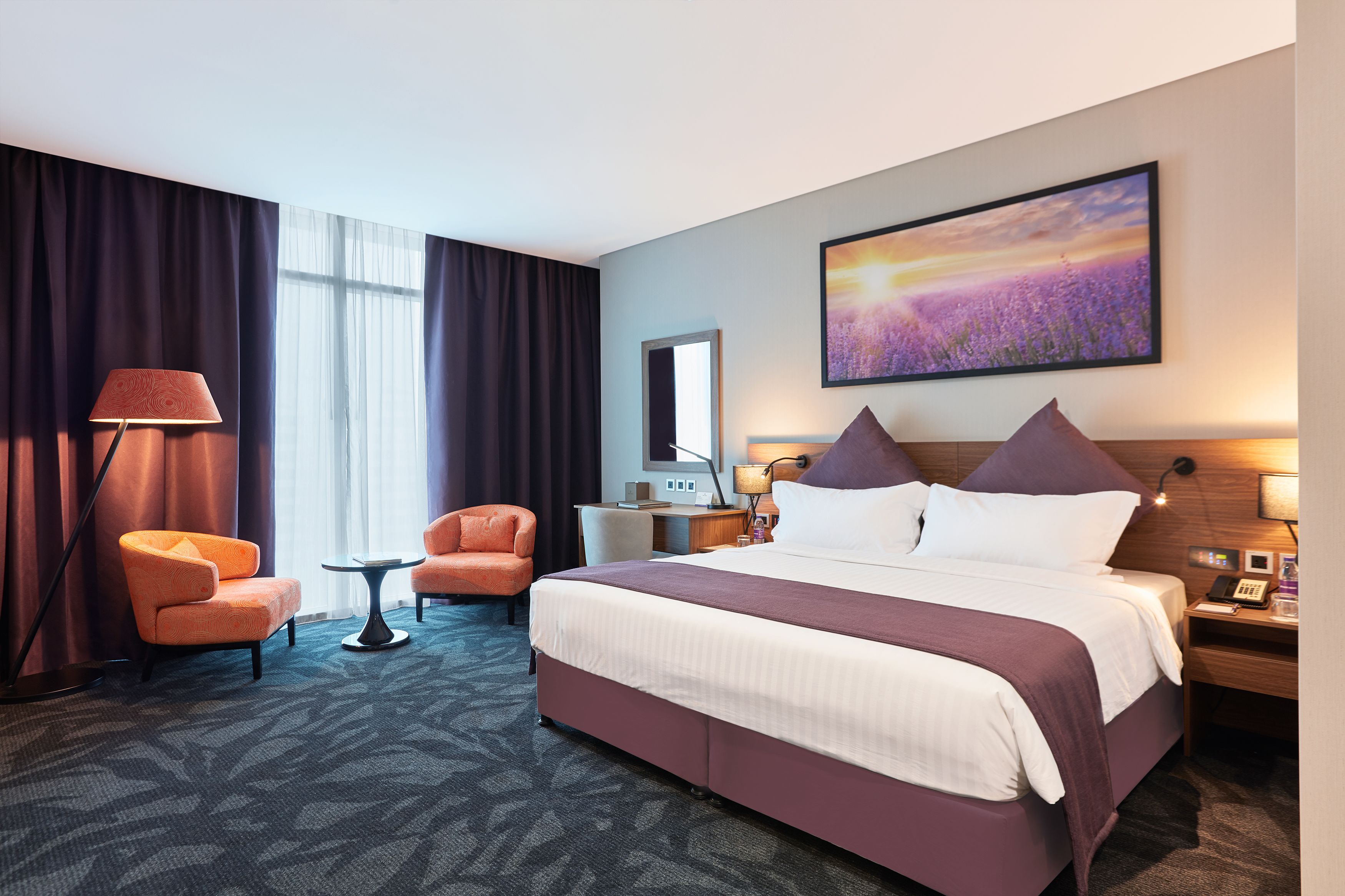 A hotel room with a large bed, two armchairs, a small table, a desk, and a large window with curtains.