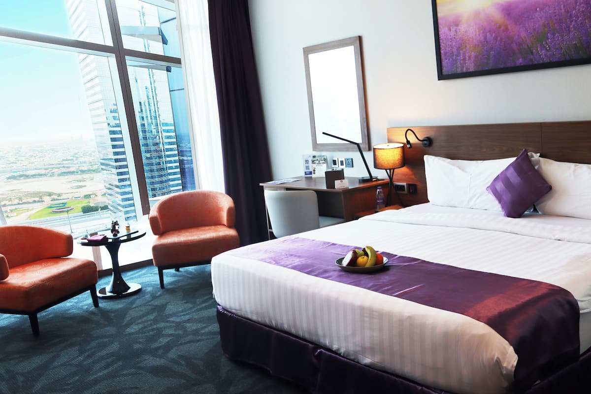 Ein Hotelzimmer mit einem großen Bett, zwei Sesseln, einem Schreibtisch mit Lampe und Blick auf eine Stadt durch große Fenster.