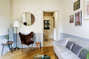 A living room with a sofa, a chair, a lamp, a mirror, and framed pictures on the wall. - Live Life Acropolis (Athens)