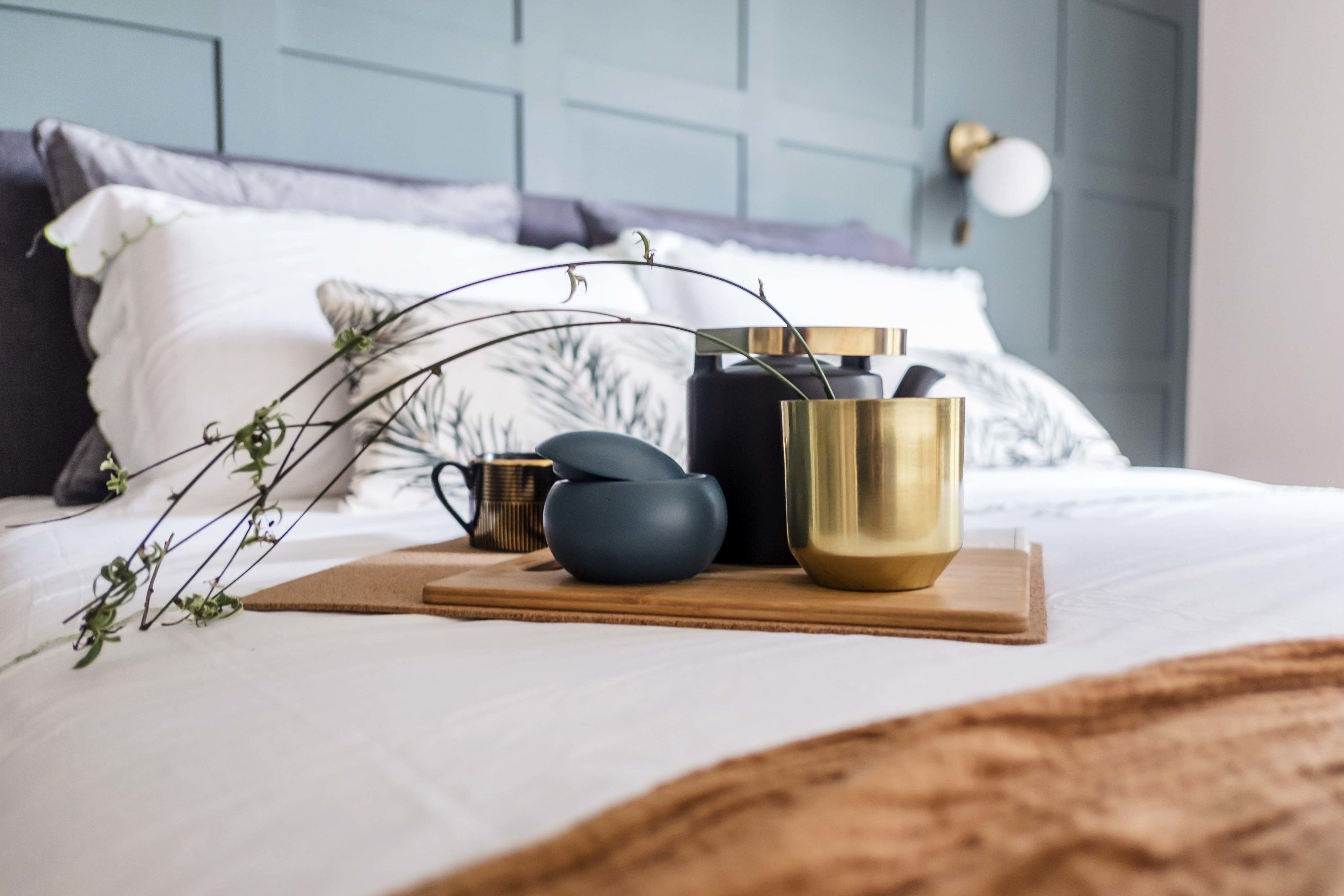A neatly arranged bed with a wooden tray holding decorative items, a blue headboard, and a wall-mounted light fixture.