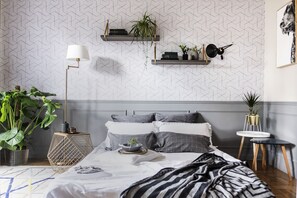 A modern bedroom with a geometric patterned wallpaper, a bed with grey and white bedding, a large green plant, a floor lamp, and two wall-mounted shelves with plants and books. - Live Life Acropolis (Athens)
