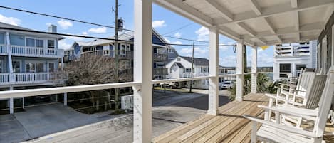 A covered porch with wooden flooring and white railings, overlooking a street with houses and parked cars.