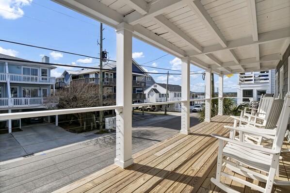 A covered porch with wooden flooring and white railings, overlooking a street with houses and parked cars.