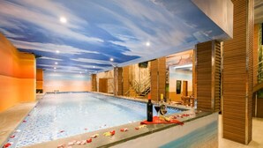 An indoor swimming pool with a sunset mural on the ceiling, a seating area with wooden panels, and rose petals on the poolside. - Caroline Hostel (Canggu)