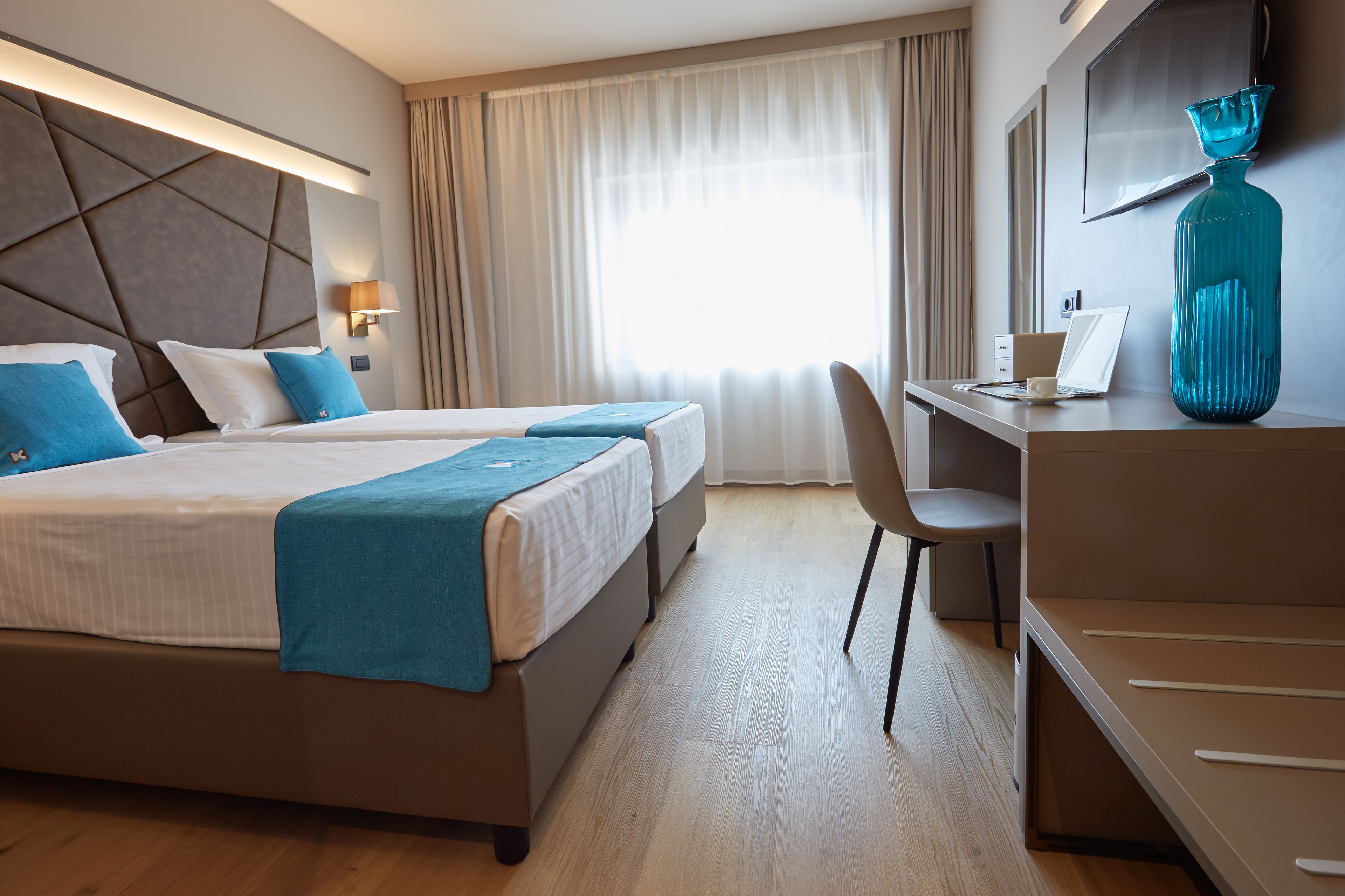 A hotel room with a large bed, a desk, a chair, a large window with curtains, and a decorative vase.