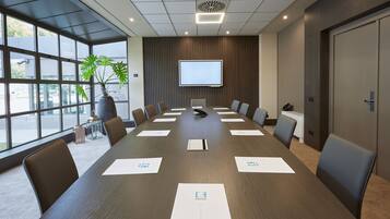 A conference room with a long table, chairs, a projector screen, and a potted plant.