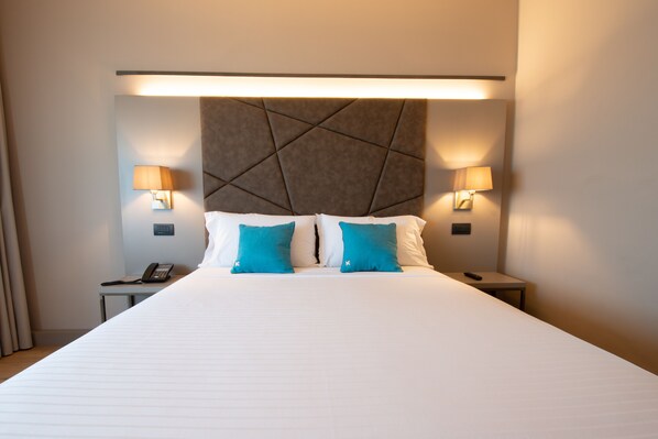 Quarto de hotel com uma cama grande, duas luminárias de cabeceira, mesa de cabeceira com telefone e cabeceira com padrão geométrico.