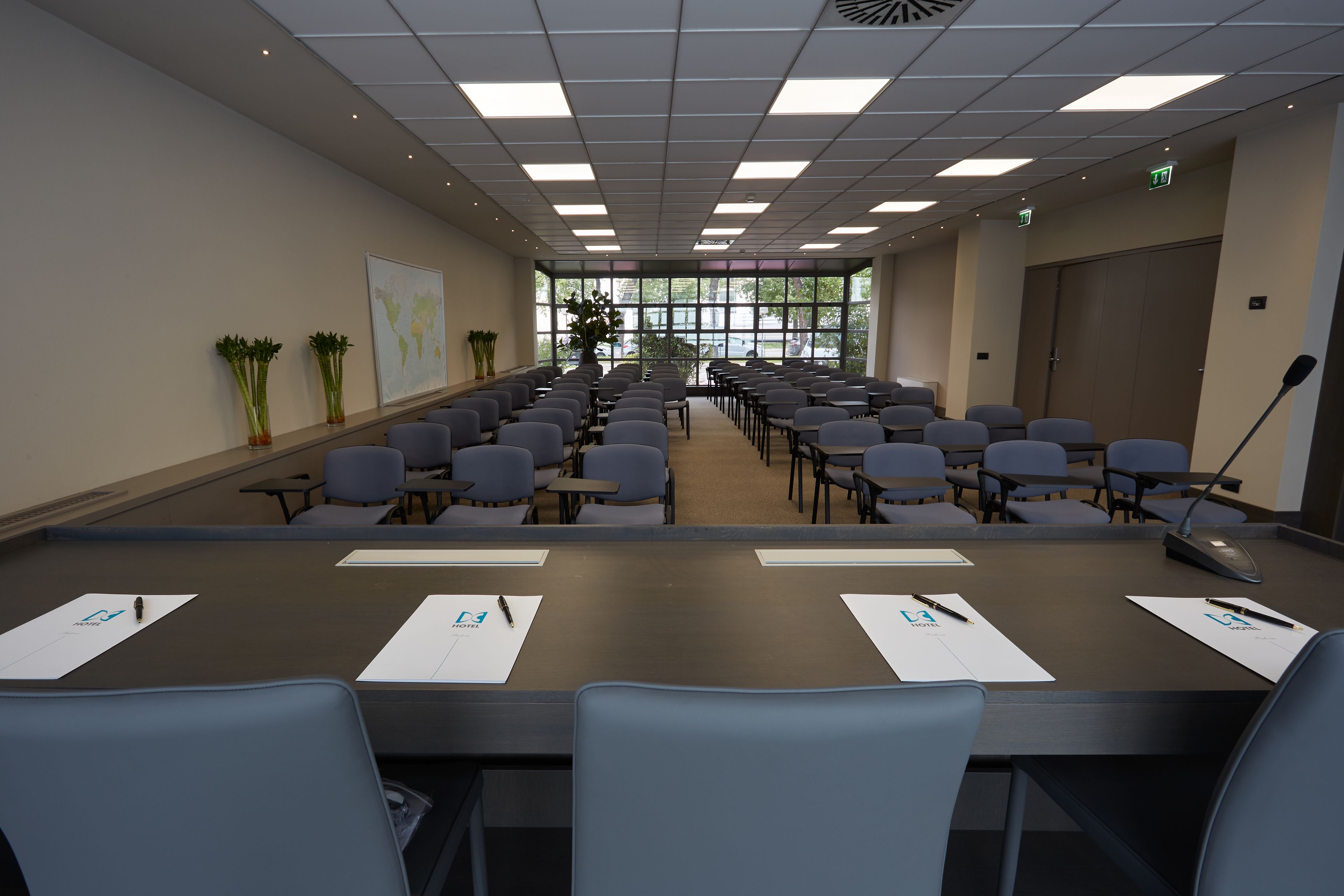 Un salón de conferencias con filas de sillas, una larga mesa con papeles y bolígrafos, y amplias ventanas que dan a vistas de árboles y un edificio.