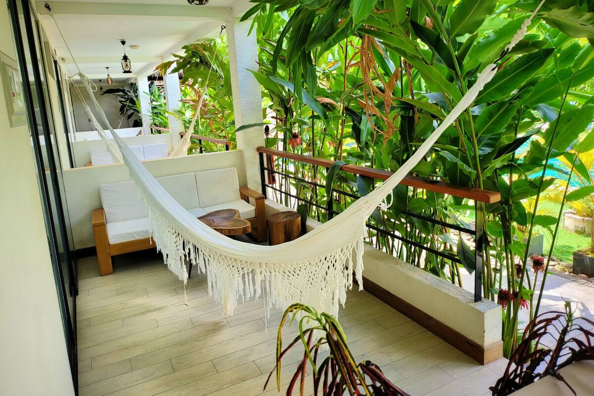 Een hangmat op een balkon met een houten vloer, omringd door weelderige groene planten en uitzicht op de omgeving.