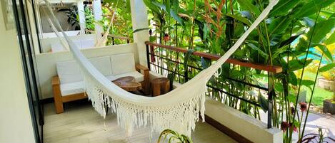 A hammock on a balcony with a wooden floor, surrounded by lush green plants and a view of the outdoors.