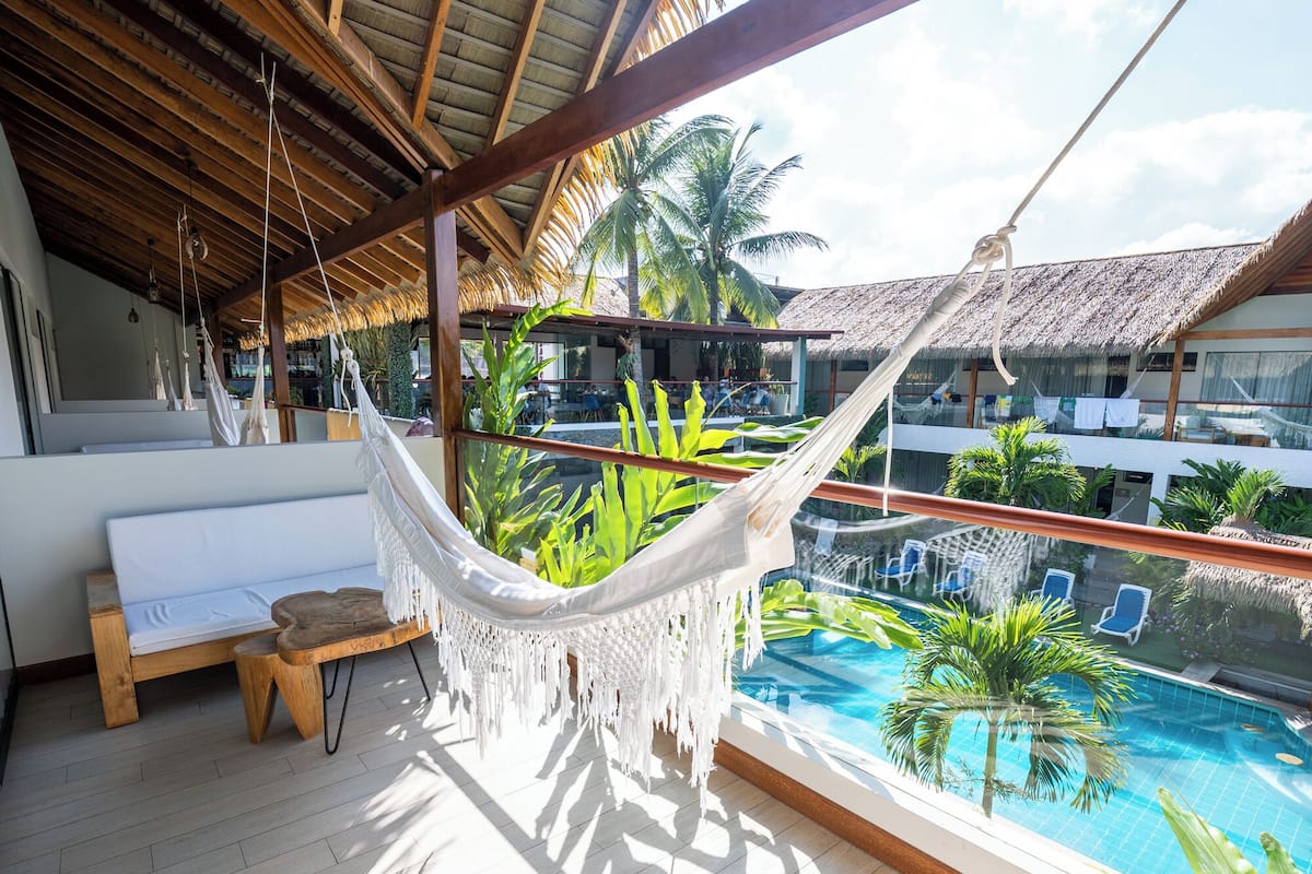 Een hangmat onder een houten plafond, een zwembad en uitzicht op een resort met palmbomen.