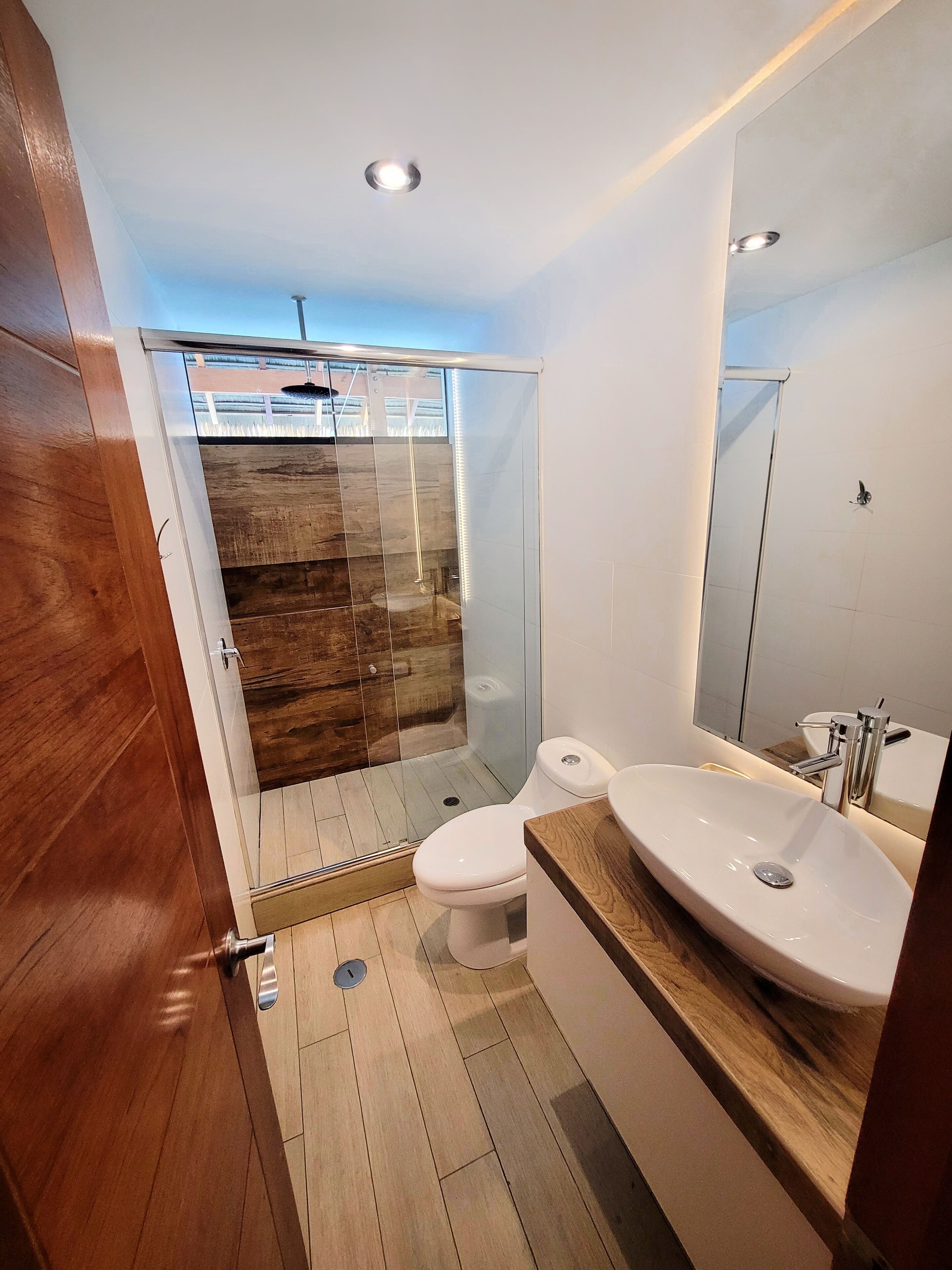 A modern bathroom with a wooden floor, a glass shower enclosure, a white toilet, and a large mirror above the sink.