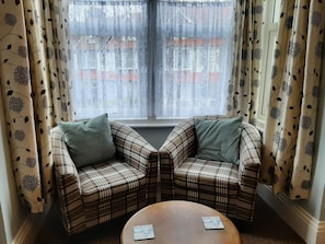 A corner seating area with two plaid sofas and a round coffee table, a window with sheer curtains, and a view of buildings outside. - Rosemead Guest house (Paignton)
