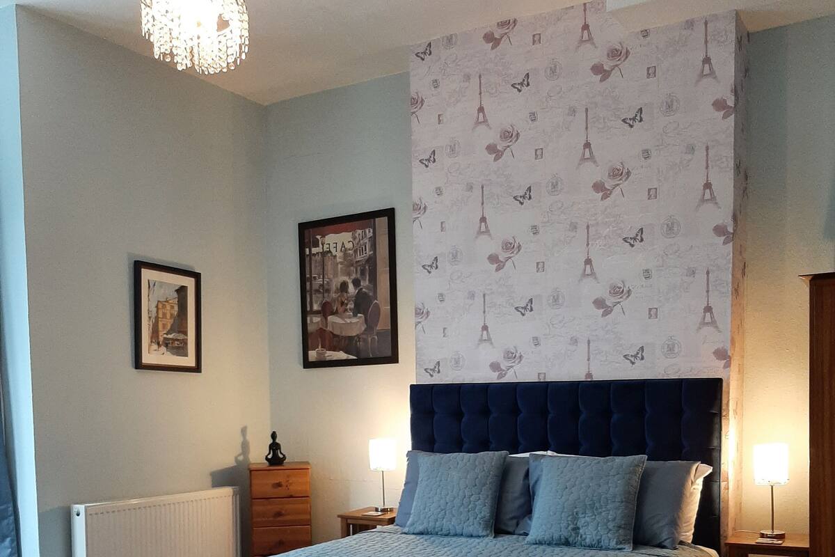 A bedroom with a large bed, a headboard with a floral pattern, a chandelier, and two bedside tables with lamps.
