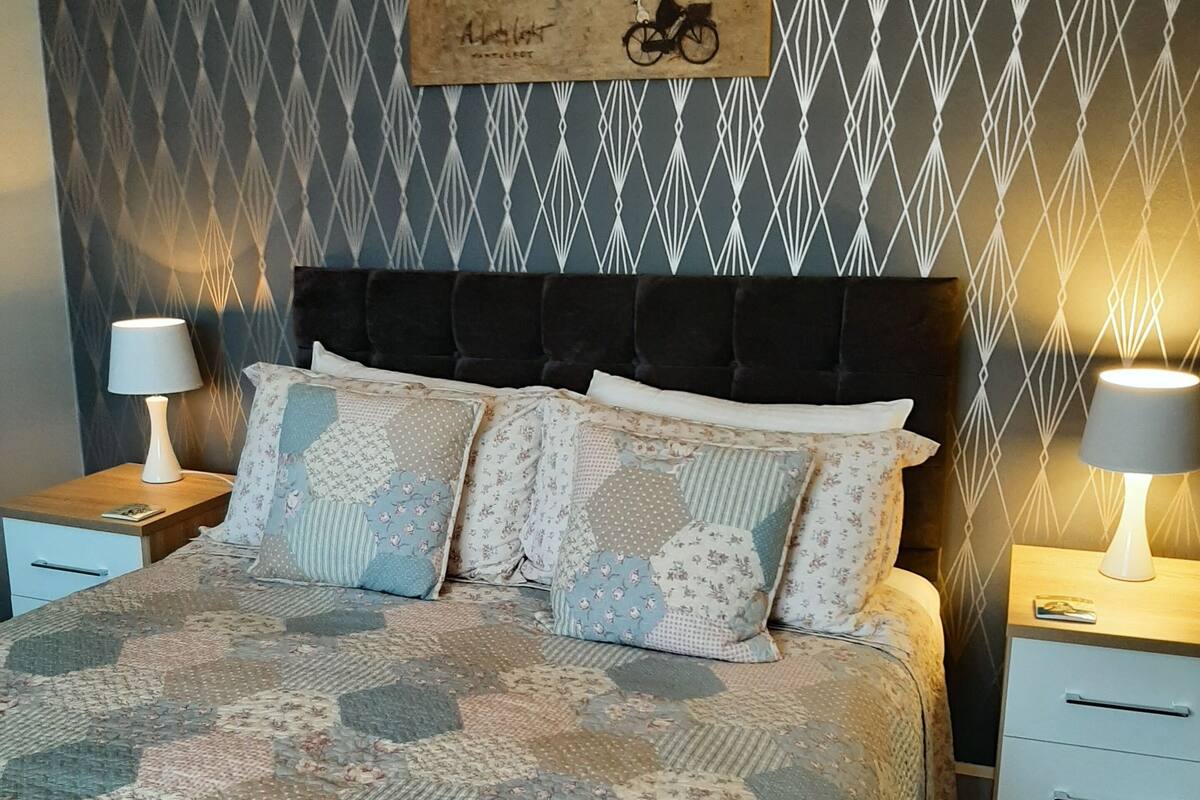 A bedroom with a bed, two bedside tables, a lamp on each table, a patterned wall, and a painting of a lighthouse.