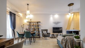 A modern living room with a sofa, armchairs, a coffee table, and a TV. There is a bookshelf with decorative items and a small table with a vase. - My Island Home (Naxos)
