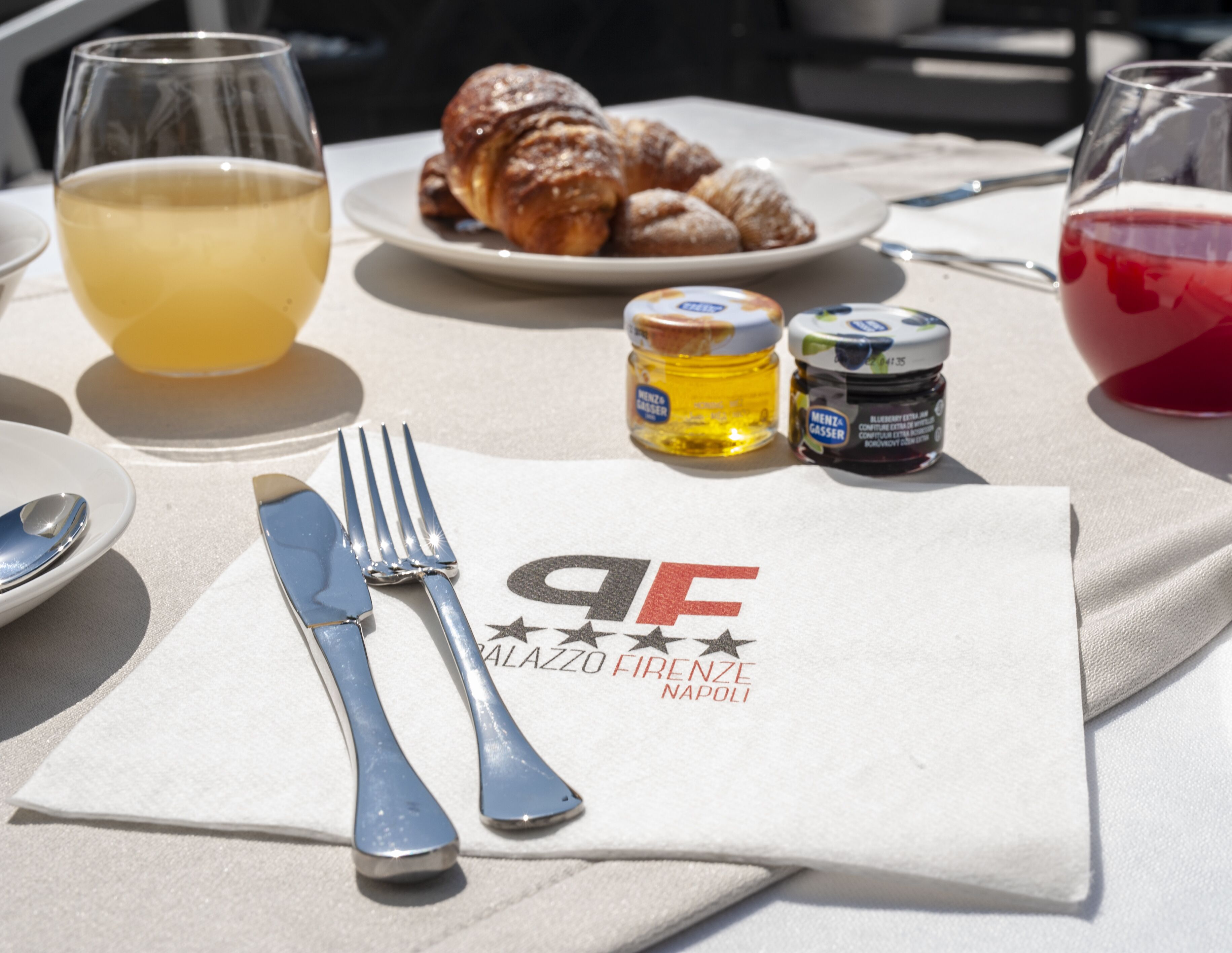 A table with a napkin, fork, knife, two glasses, a plate with croissants, and two jars of jam.