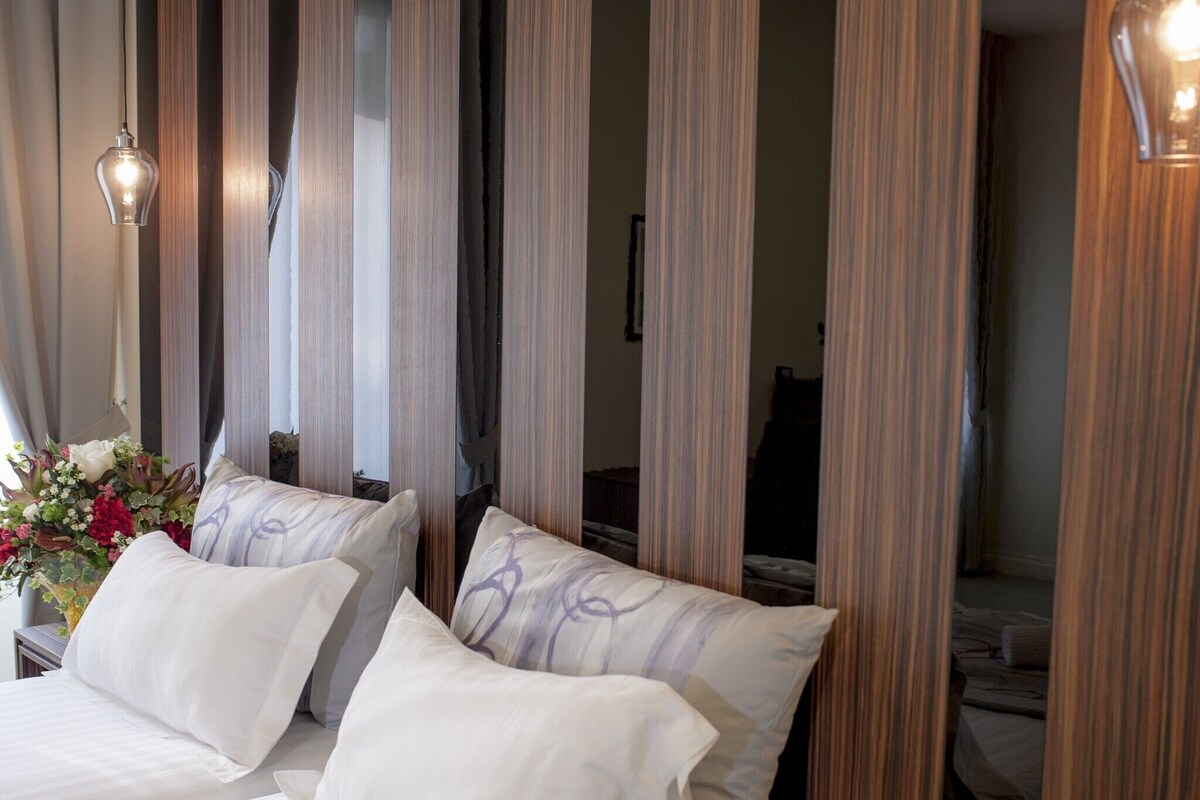 A hotel room with a bed, curtains, a nightstand with a vase of flowers, and a visible power outlet.