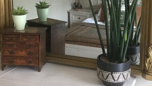 A bedroom with a large mirror, a bed with a patterned bedspread, two potted plants, a nightstand with a lamp, and a wall tapestry. - Bambu Cottage (Newquay)