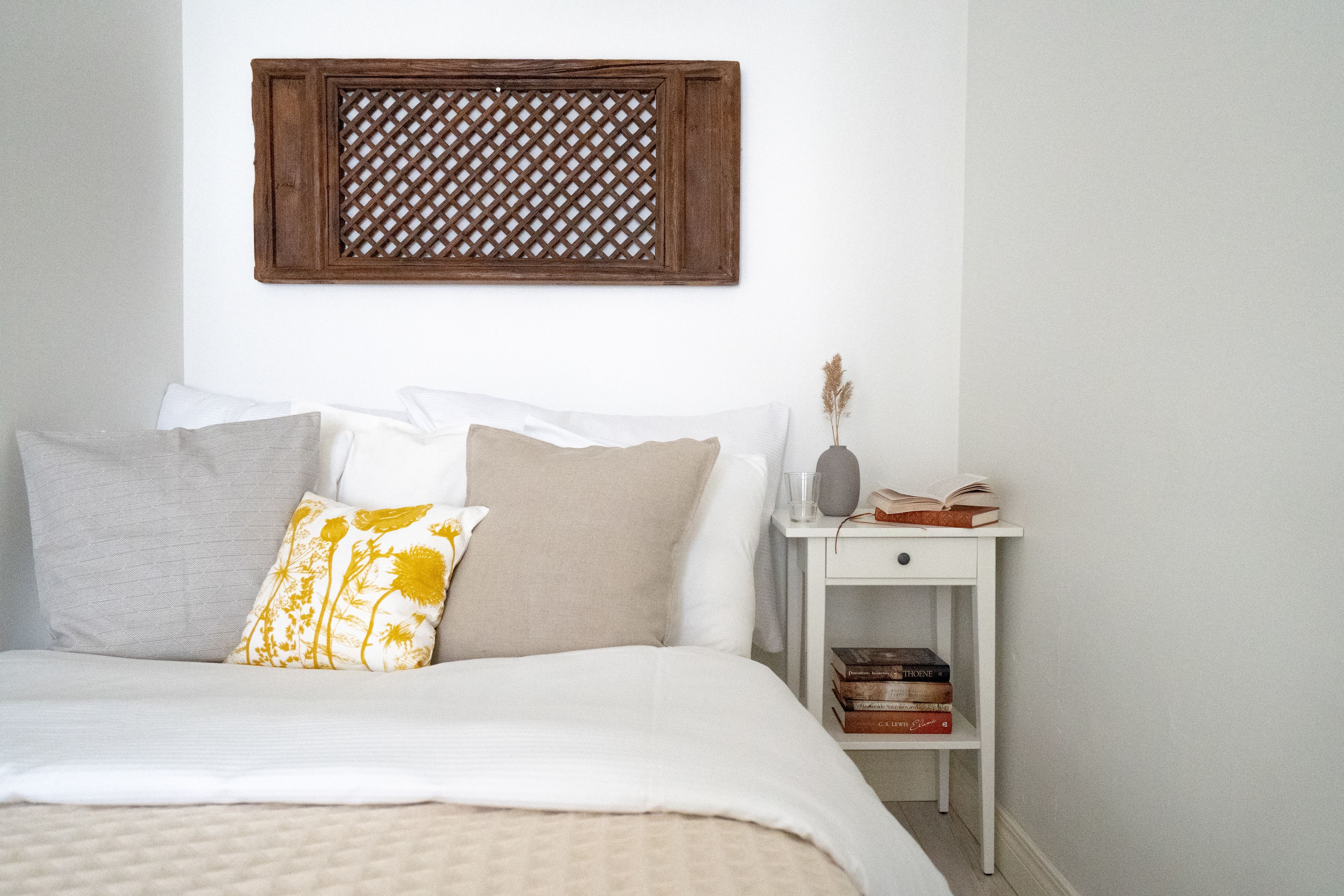 A neatly made bed with a white comforter and multiple pillows, a small bedside table with books and a vase, and a wooden lattice wall decoration.