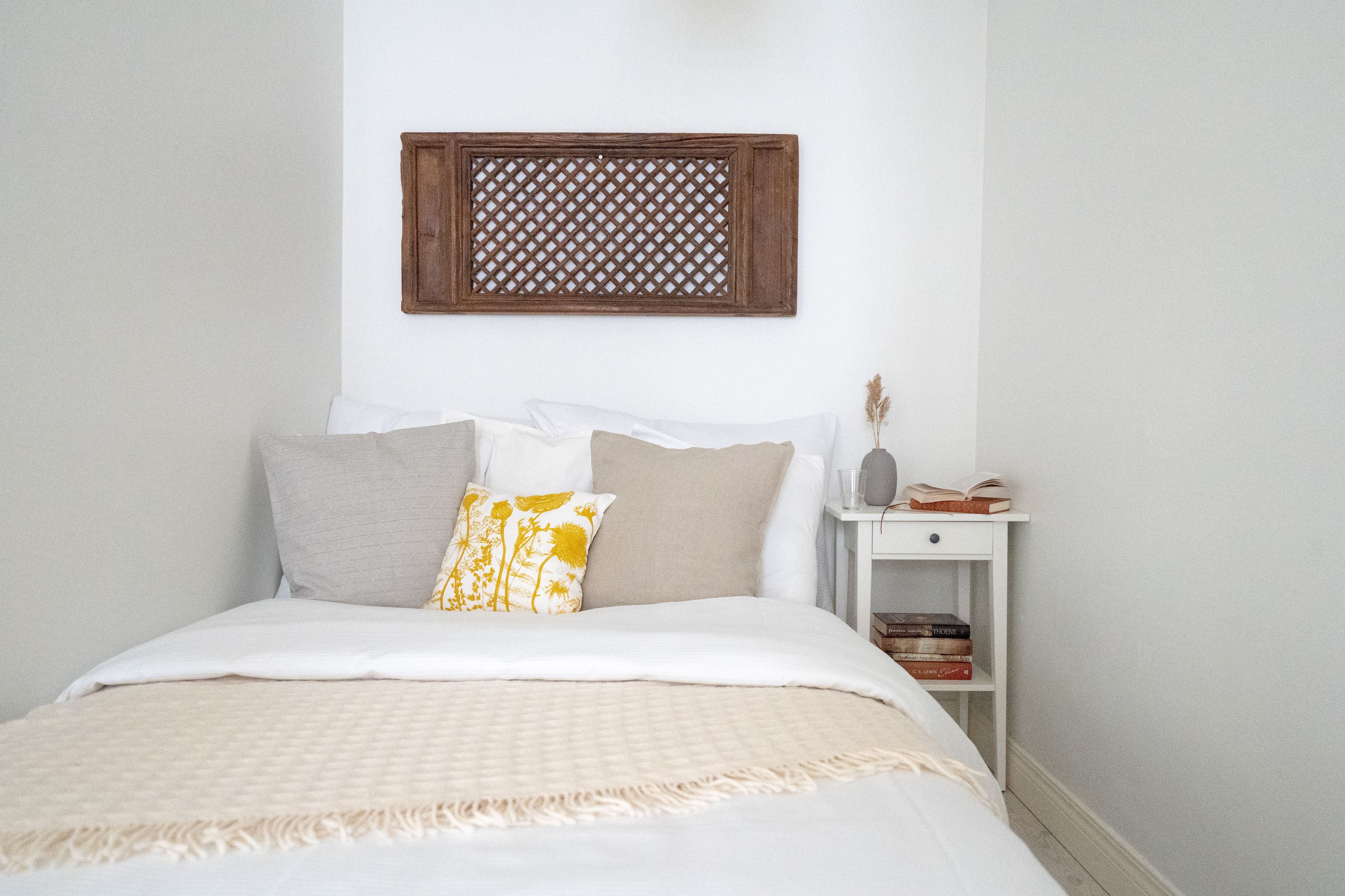 A neatly made bed with a white and beige bedspread, a wooden lattice wall decoration above it, and a bedside table with books and a vase.