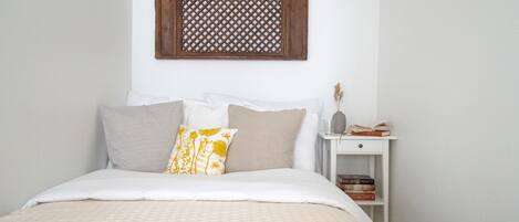 A neatly made bed with a white and beige bedspread, a wooden lattice wall decoration above it, and a bedside table with books and a vase.