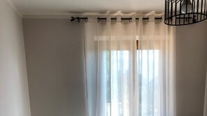 Un lit avec une couvre-lit matelassée blanche et beige, une tête de lit en métal noir et des rideaux transparents qui laissent passer la lumière naturelle.