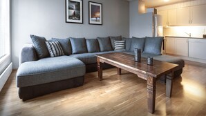 Una sala de estar moderna con un sofá modular, una mesa de centro de madera y una cocina al fondo.