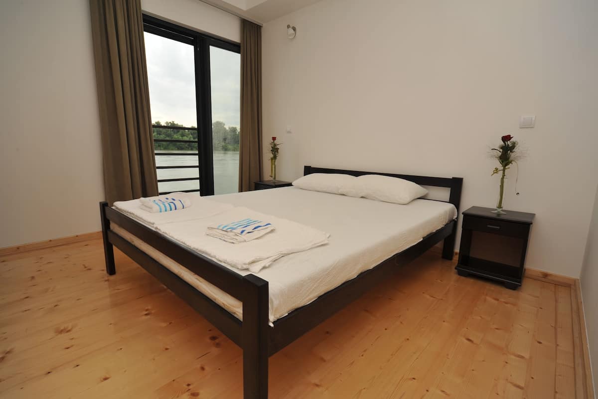 A bedroom with a wooden floor, a bed with white bedding, a nightstand, and a window with curtains.