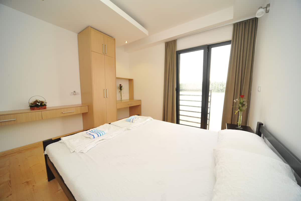 A hotel room with a large bed, a wooden wardrobe, a small table with a vase, and a view through a sliding door.