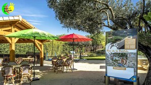 An outdoor seating area with wicker tables and chairs, covered by green and red umbrellas, and a signboard with information. - Mas de la Bastide (Joyeuse)