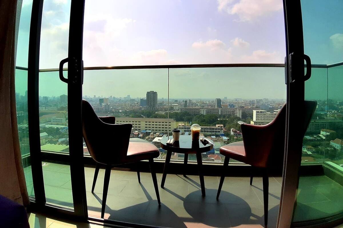 A balcony with two chairs and a table, overlooking a cityscape with buildings and greenery.