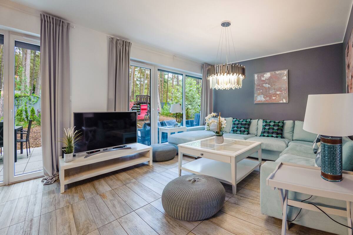 A modern living room with a flat-screen TV, a sofa, a coffee table, and a large window offering a view of the outdoors.