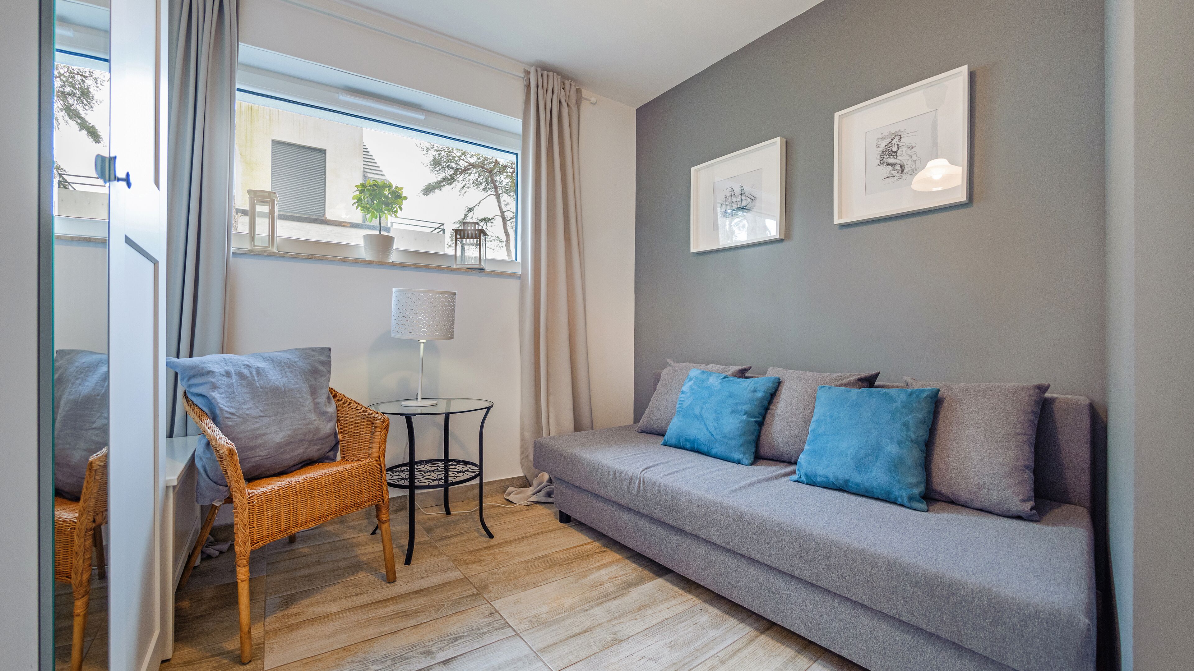 A modern living room with a grey sofa, blue cushions, a wicker chair, a small table with a lamp, and a large window with curtains.