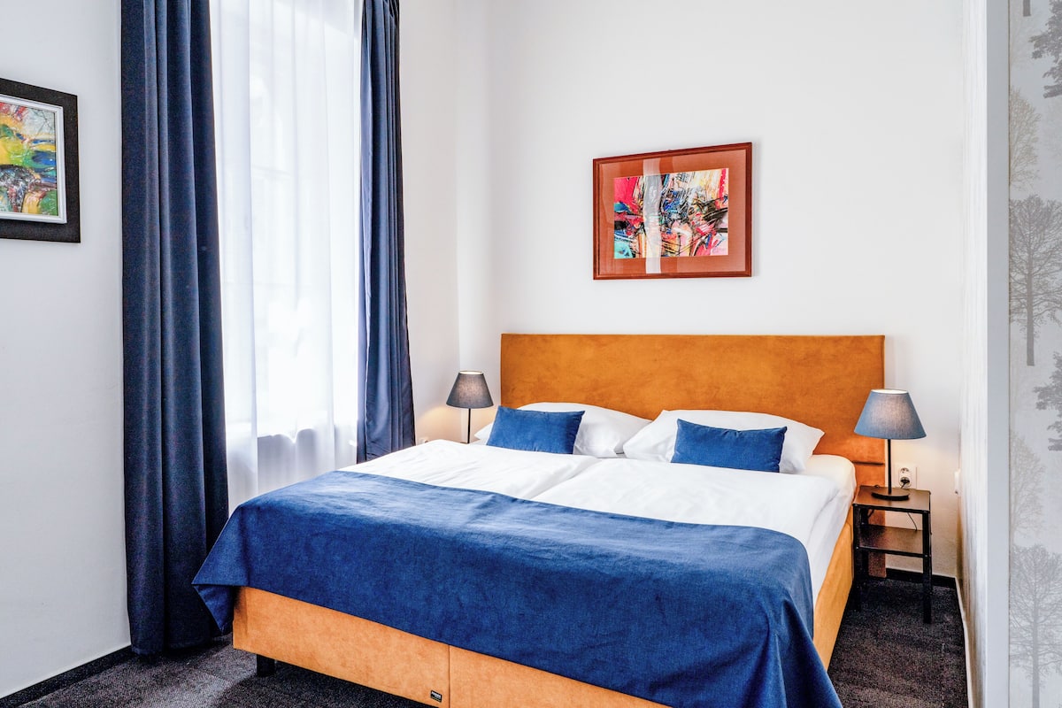 A hotel room with a large bed, two bedside tables, a nightstand, a window with curtains, and a framed picture on the wall.