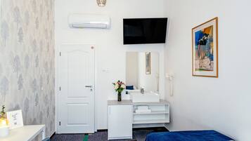 Hotelkamer met een witte deur, een flatscreen televisie aan de muur, een witte kaptafel met spiegel, een bed met een blauwe deken en een schilderij.