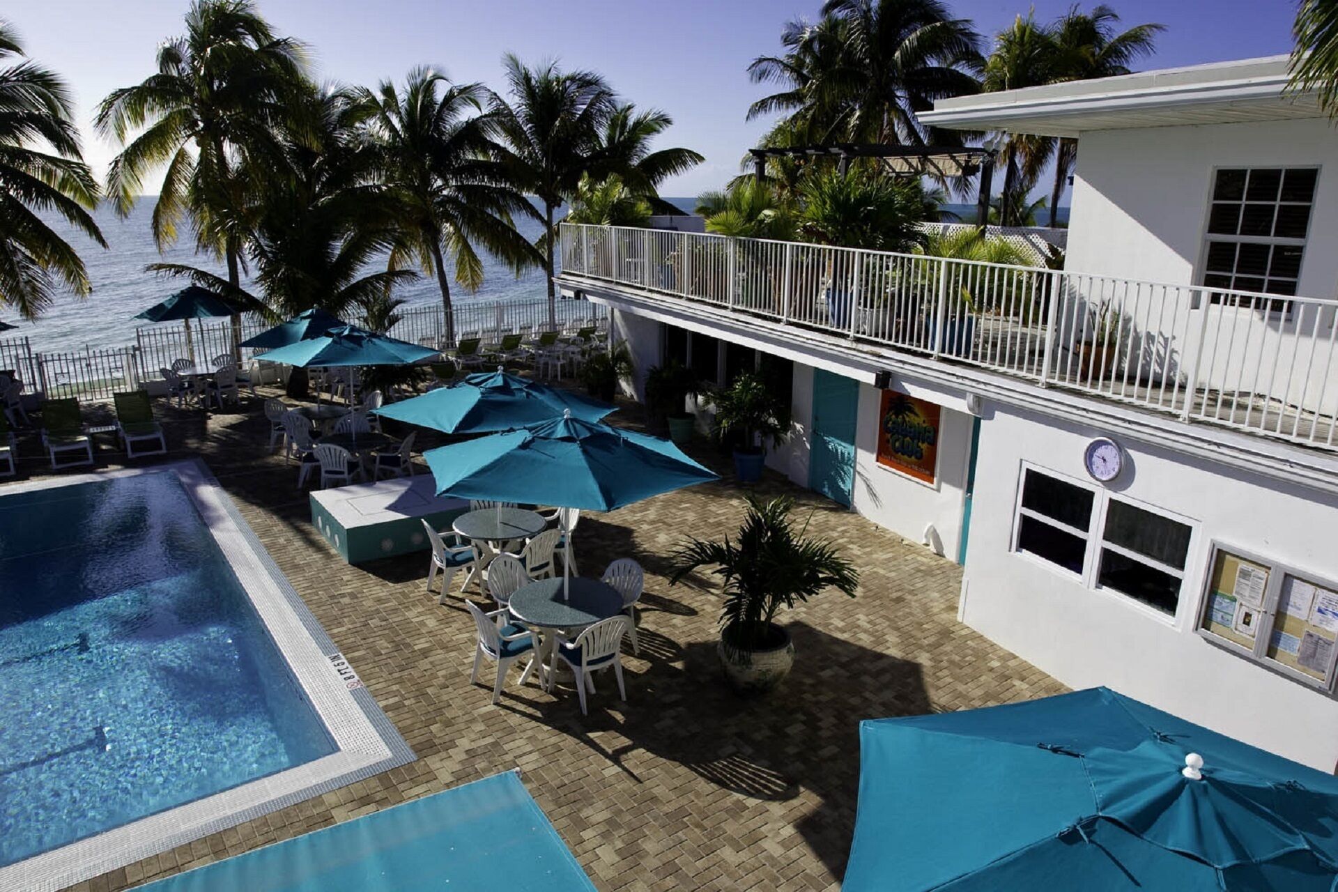 A pool area with lounge chairs and umbrellas, a building with a balcony, and palm trees in the background.