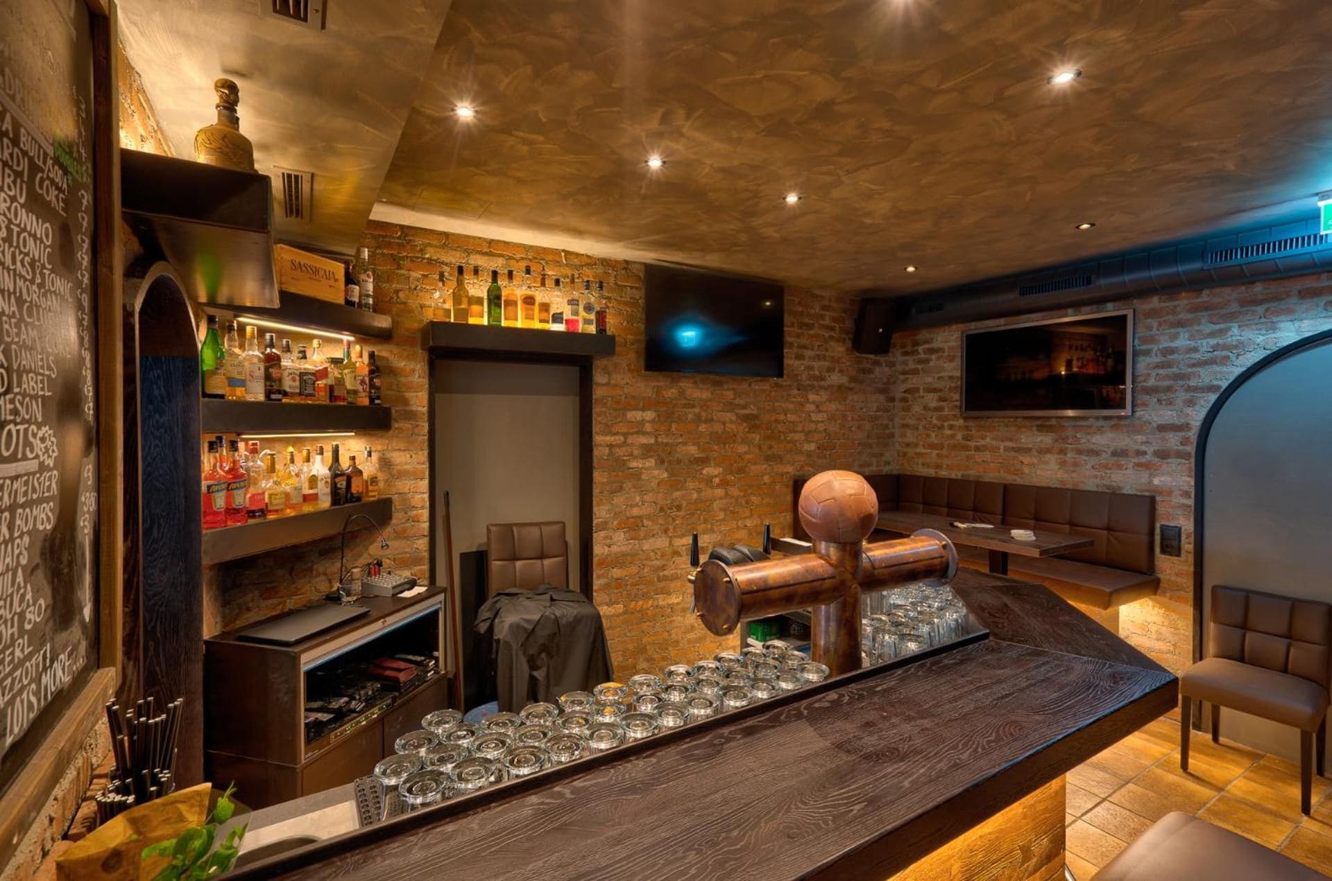 A bar area with a brick wall, shelves of liquor bottles, a billiard table, and a seating area with a television screen.