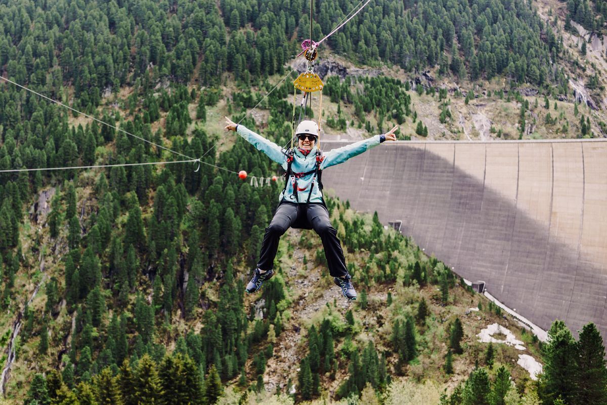 A person suspended in mid-air with outstretched arms, wearing a helmet and harness, against a backdrop of a forested mountain and a large dam.