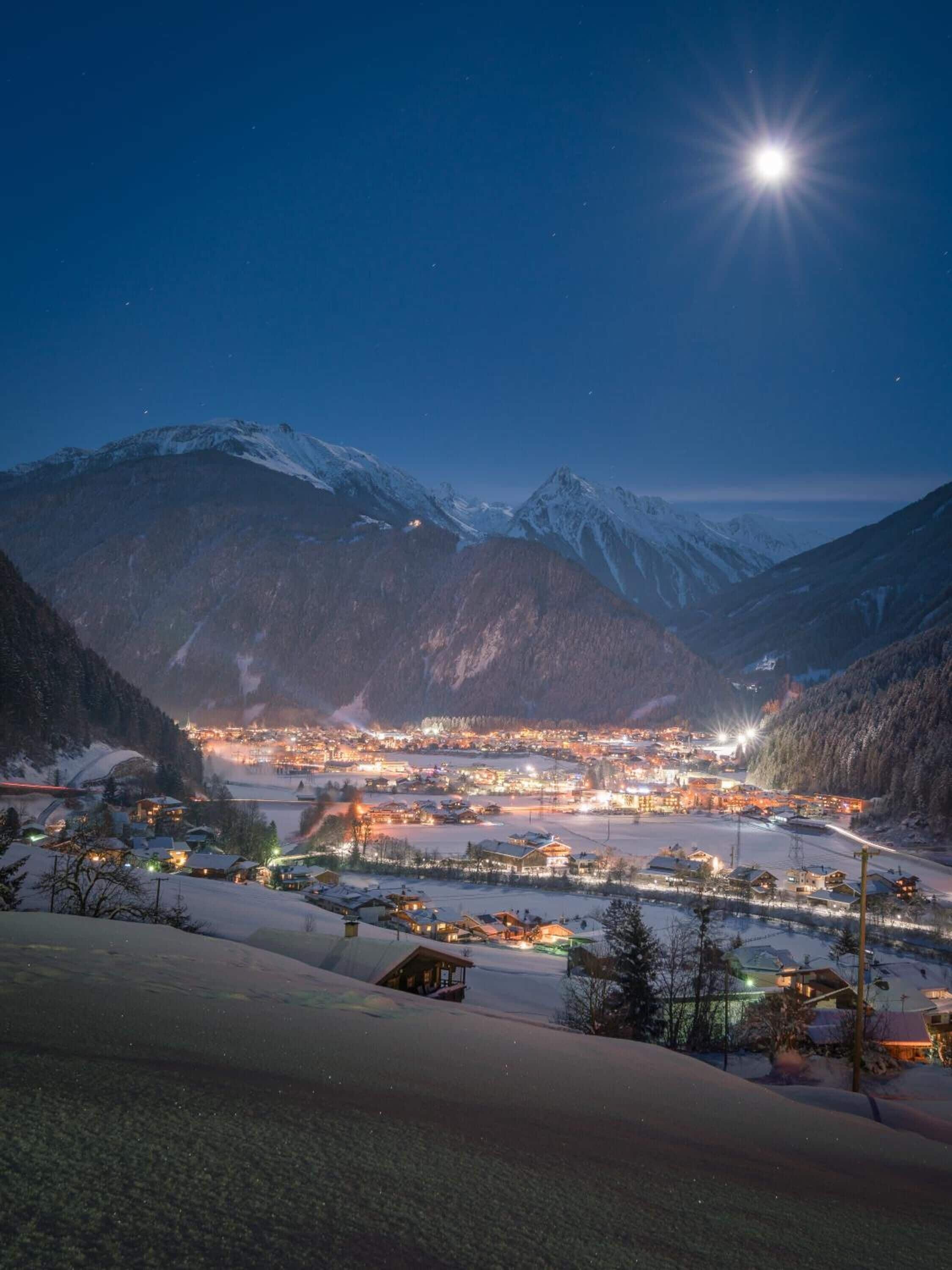 Un pueblo cubierto de nieve al caer la noche, con una luna brillante y estrellas en el cielo.