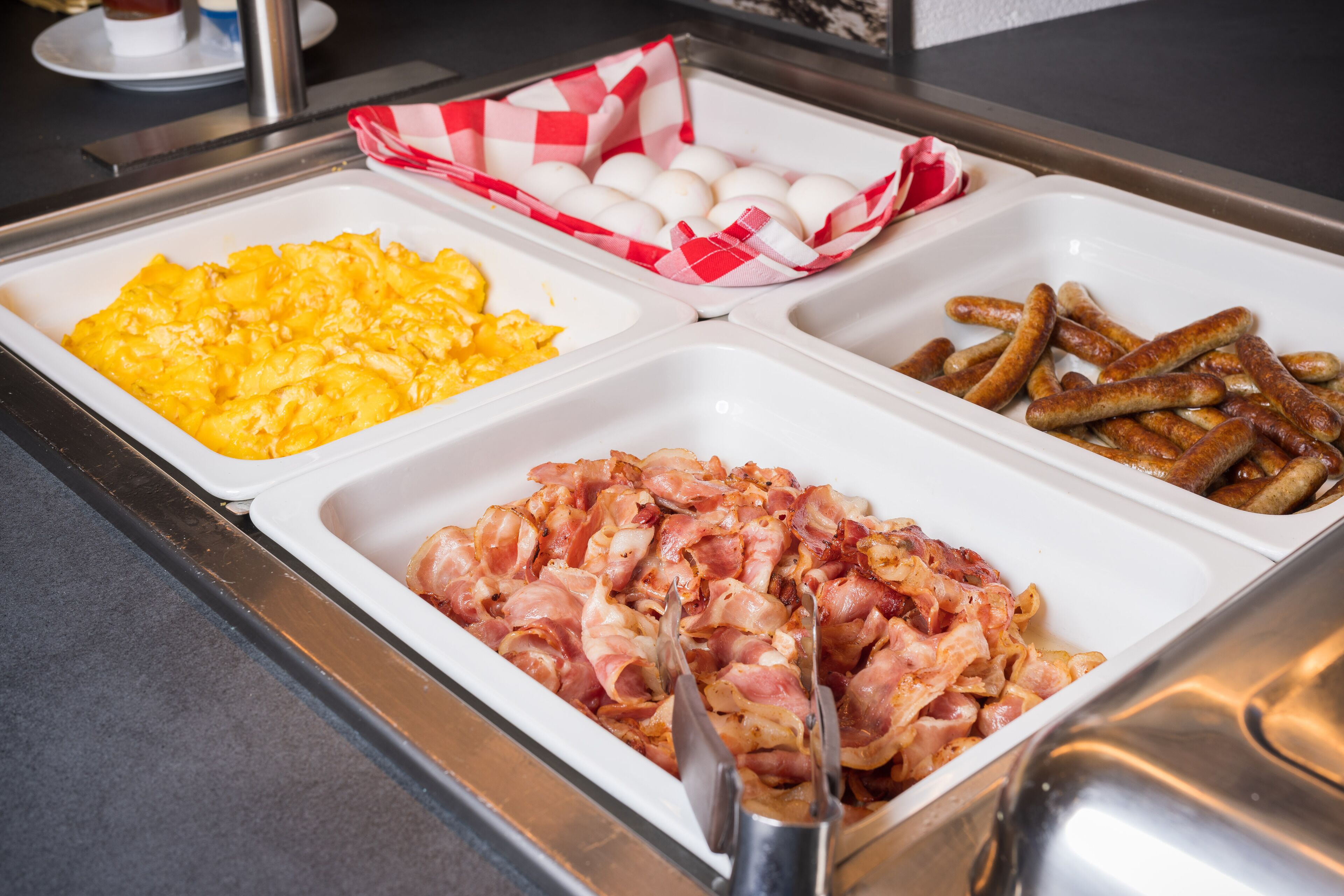 Buffet com ovos mexidos, bacon e salsichas.