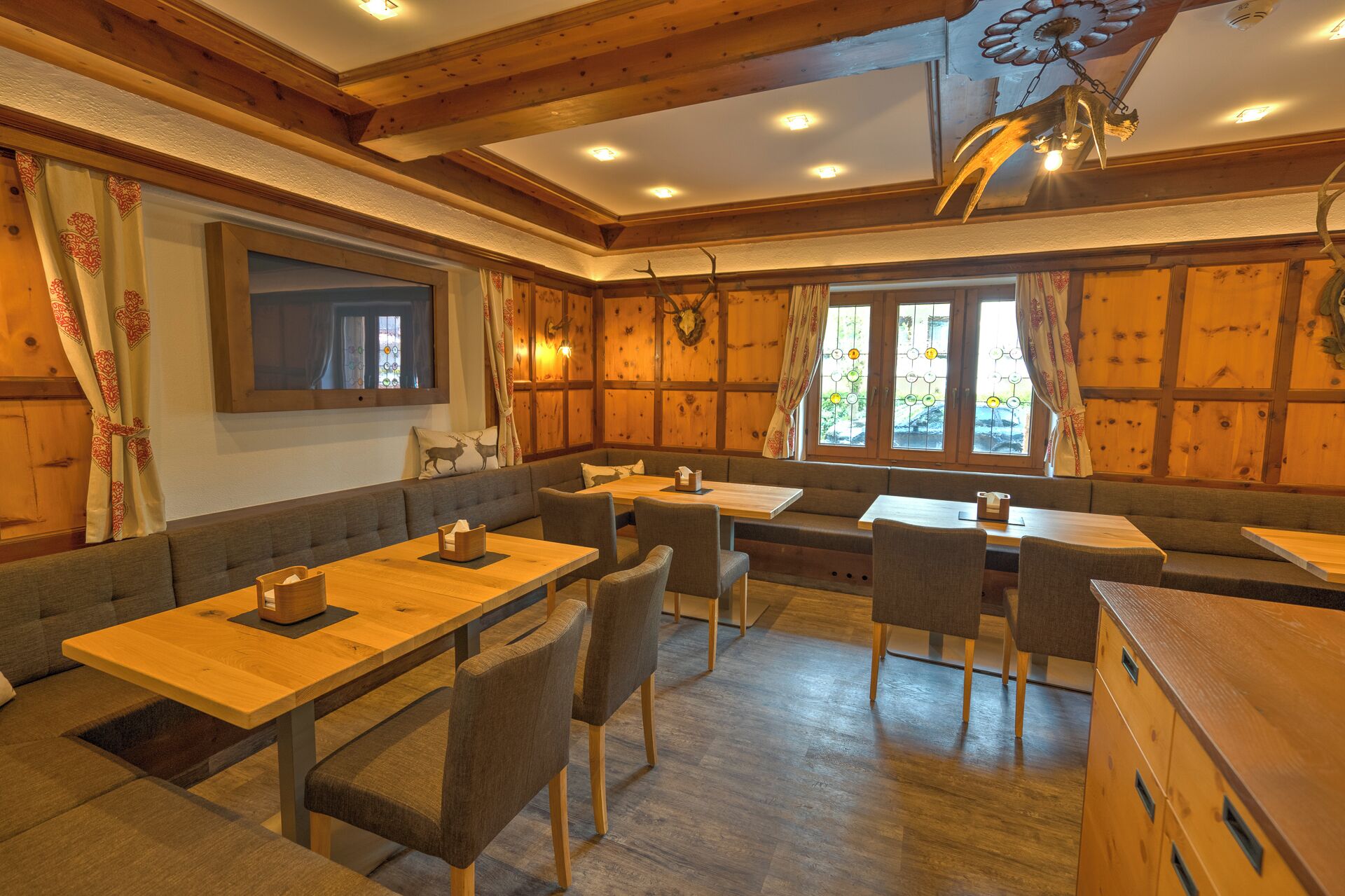 A cozy restaurant with wooden walls and ceiling, featuring a large wooden table and benches, and a decorative antler hanging from the ceiling.