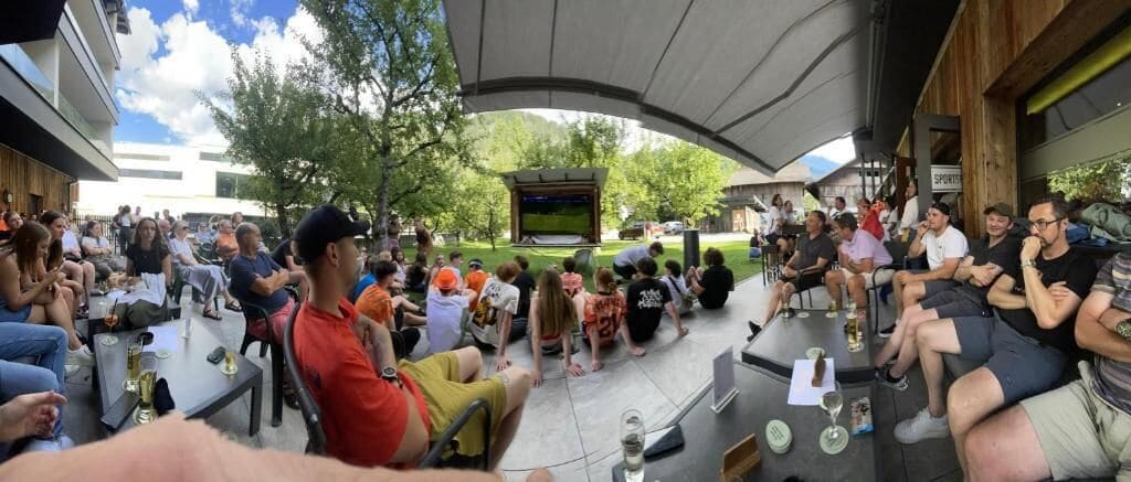 Une réunion en plein air où les participants sont assis à des tables et sur des chaises, avec une scène en arrière-plan et un espace abrité offrant de l’ombre.