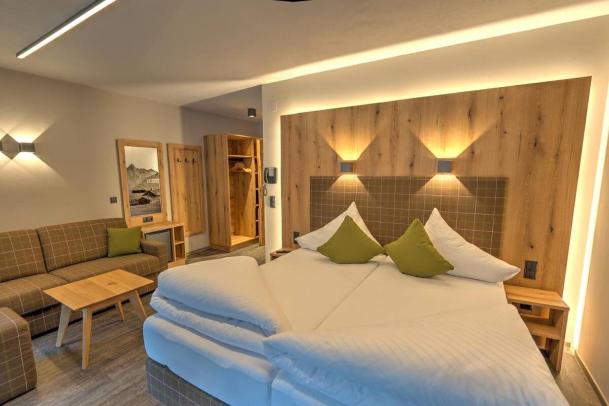 A modern hotel room with a large bed, a sofa, a small table, and wooden headboard lighting.