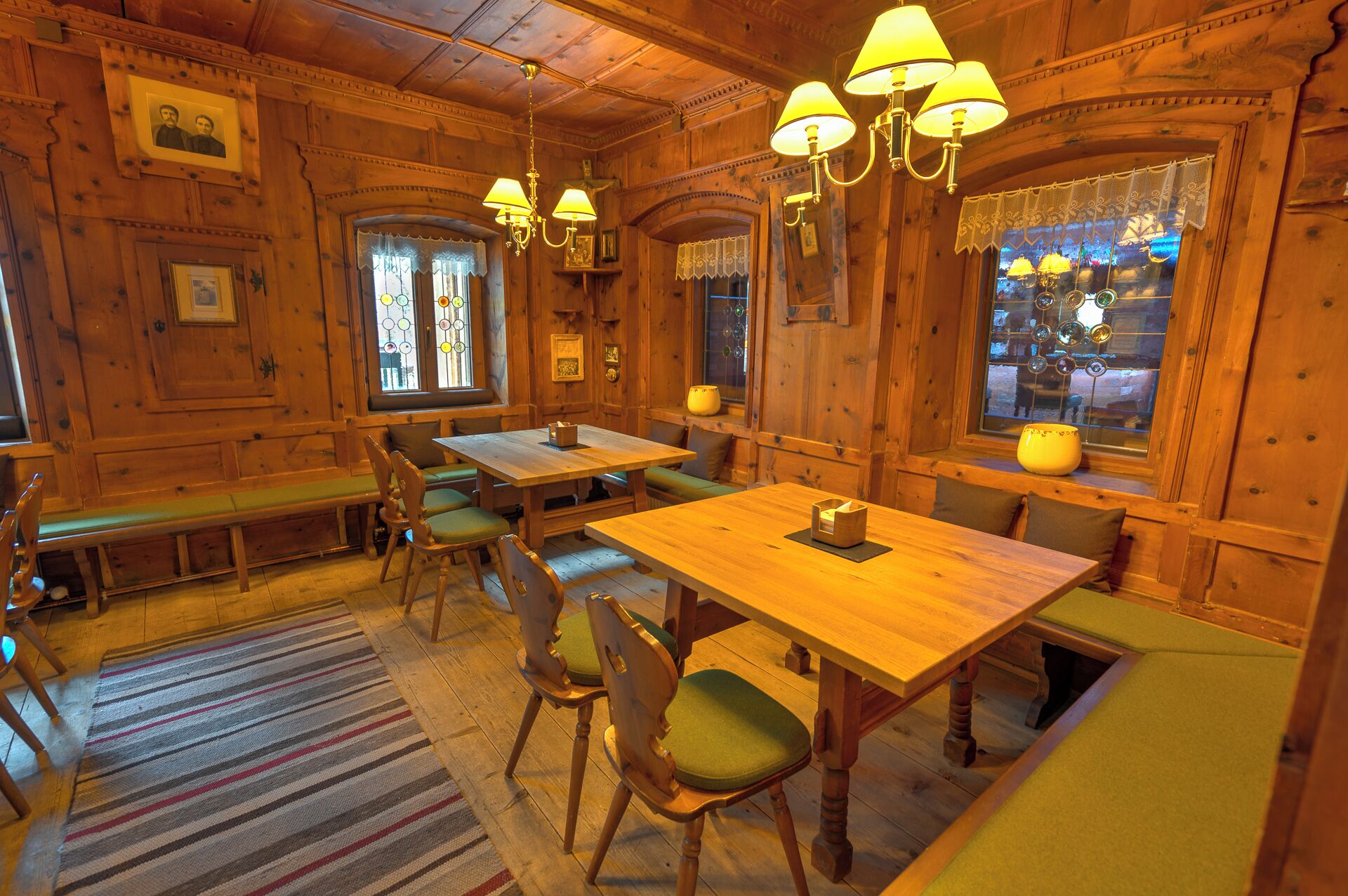 A cozy wooden interior with a dining area featuring benches and tables, a striped rug, and decorative lamps.